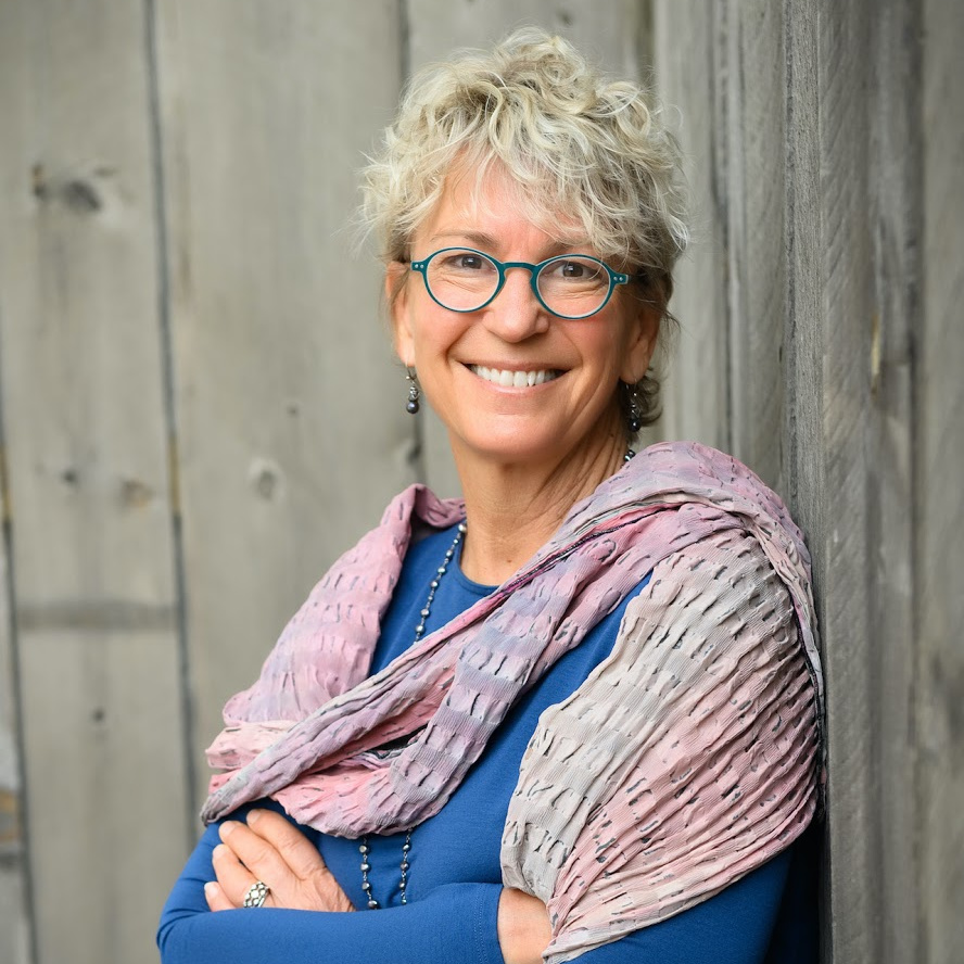 Smiling person with short blonde hair, wearing glasses and a pastel-colored shawl.
