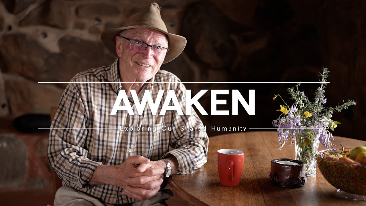 An older man sits at a table with his hands clasped and a smile on his face. Text that reads "Awaken" overlays the image.