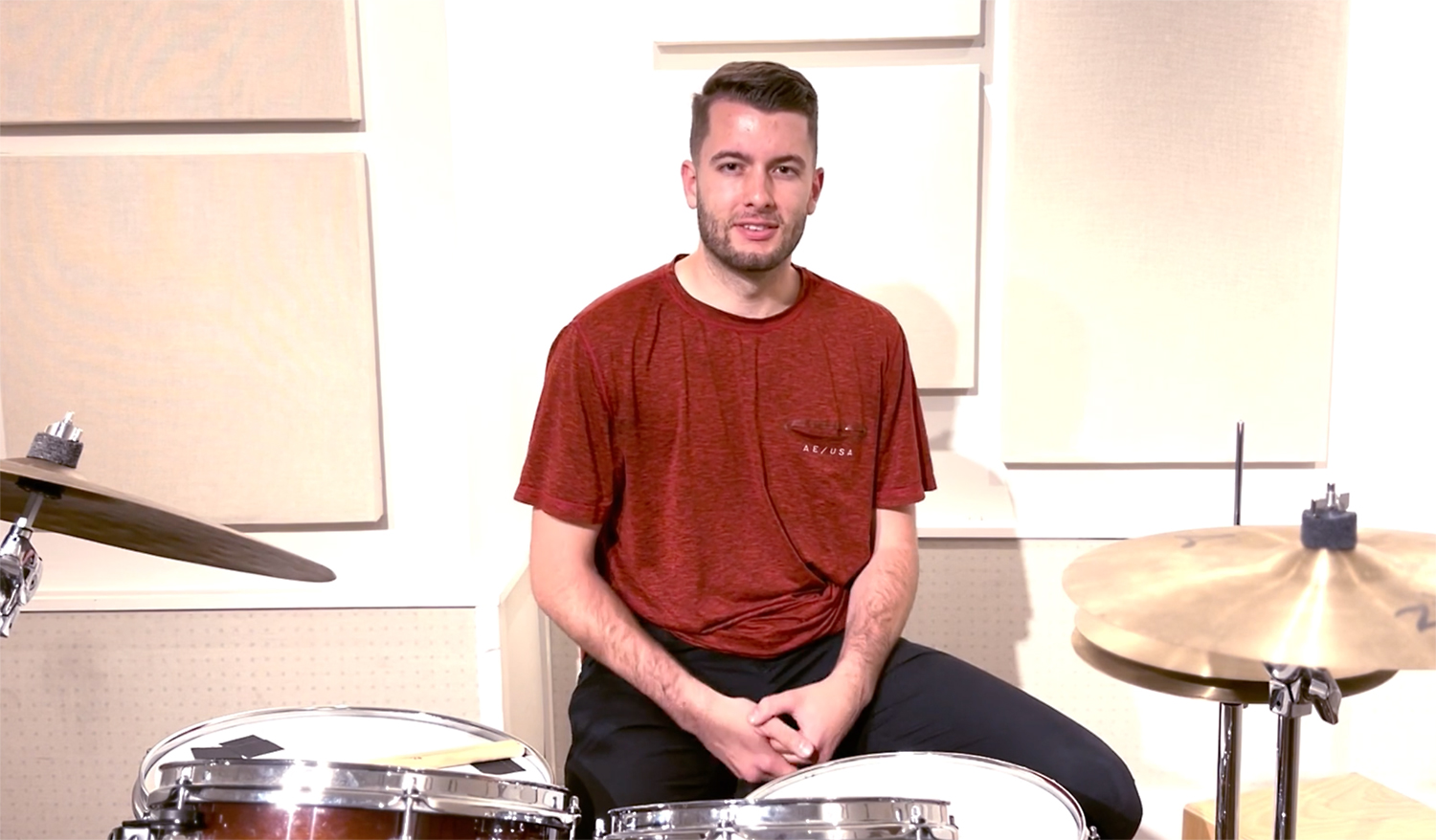 Young white man in a red t-shirt sitting in front of a drum set against a white background