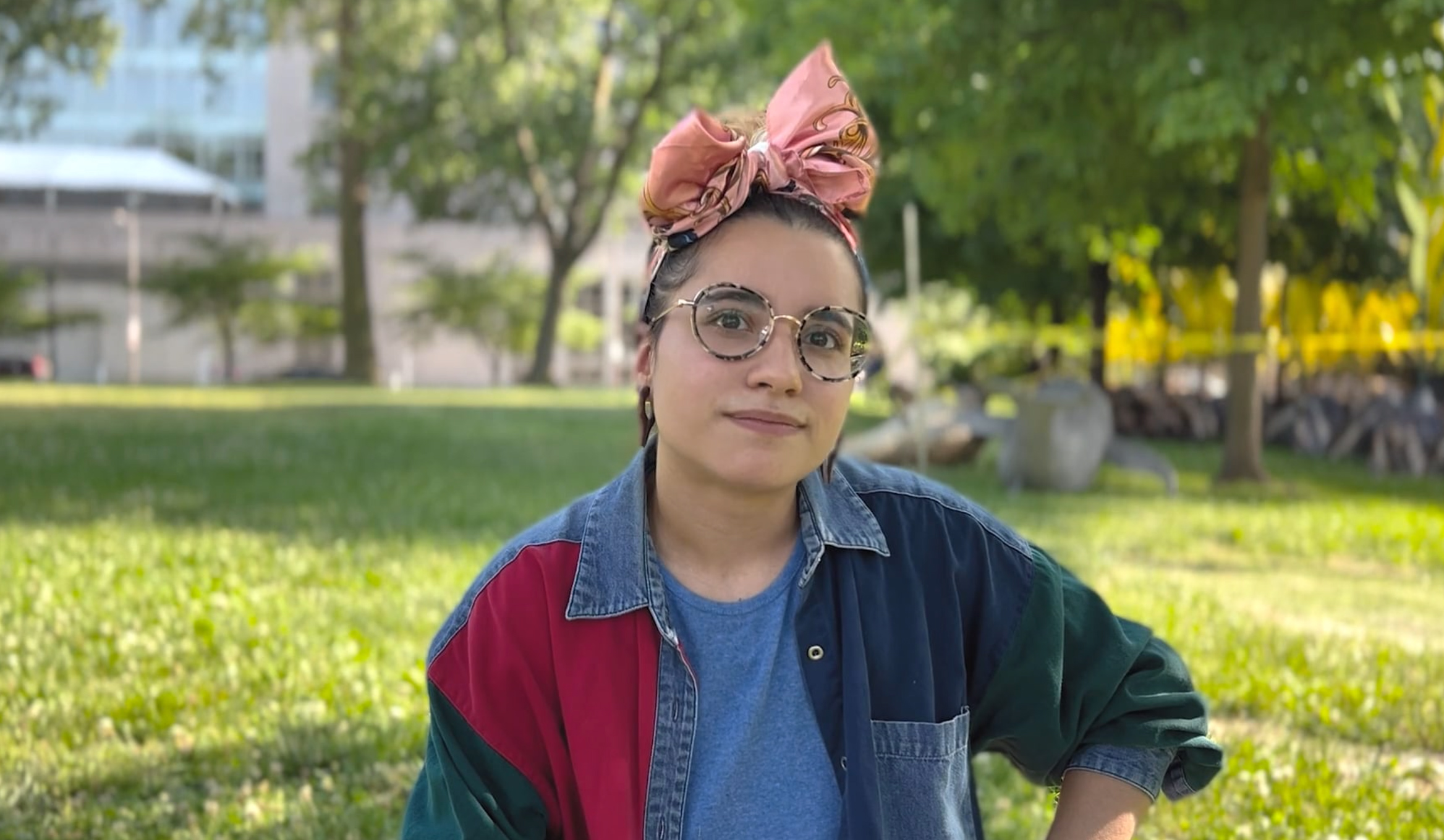 A young woman wearing glasses sitting in a grassy park. The grass is speckled with sunshine, and trees and a tan building are in the background.