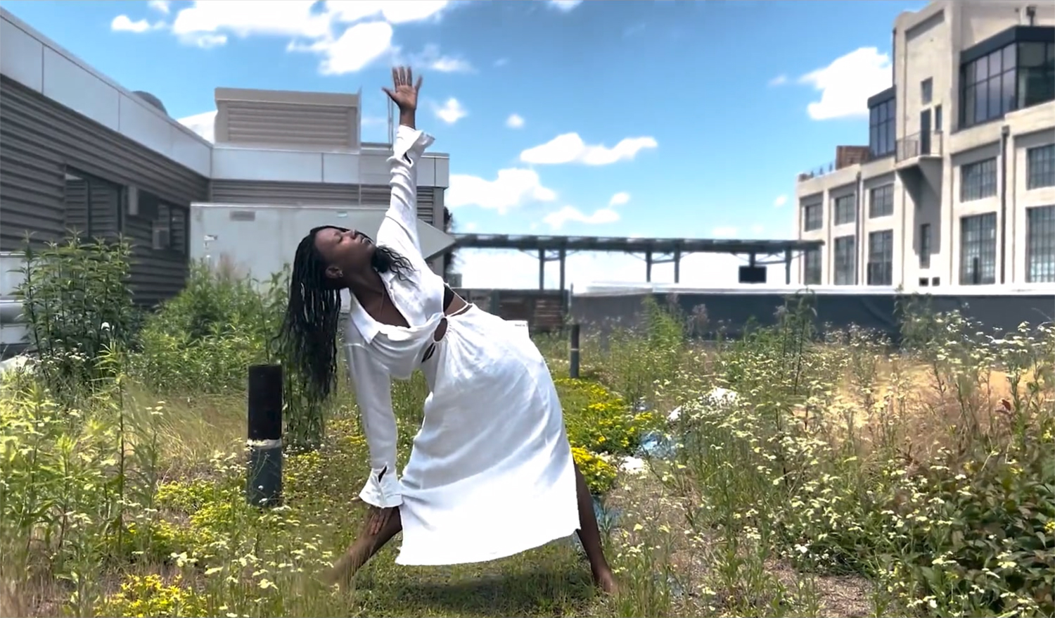 A young woman in a long white dress practicing yoga in an overgrown field between two industrial buildings.