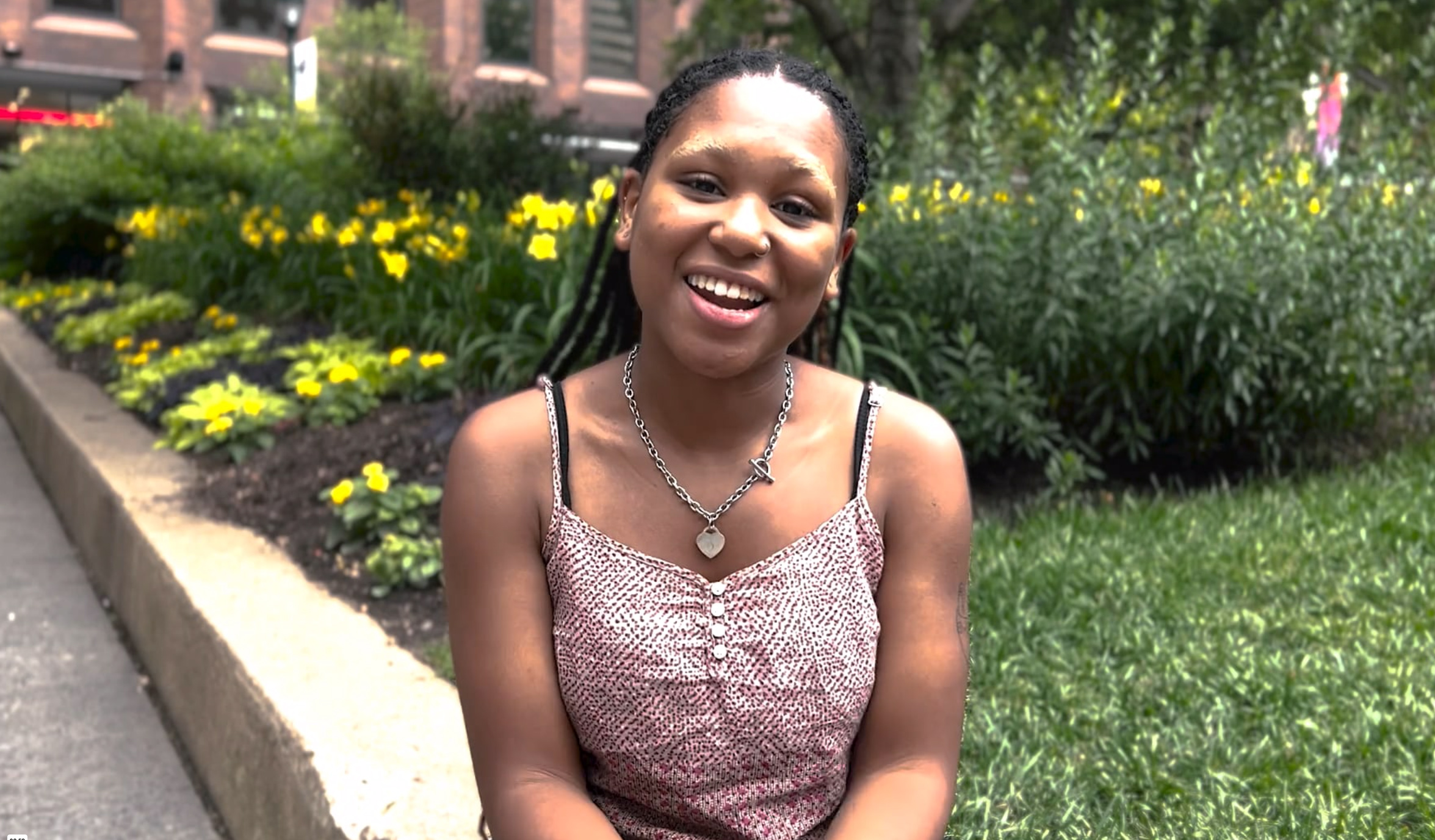 A young woman sitting on a city sidewalk ledge in front of green grass and yellow daffodils.