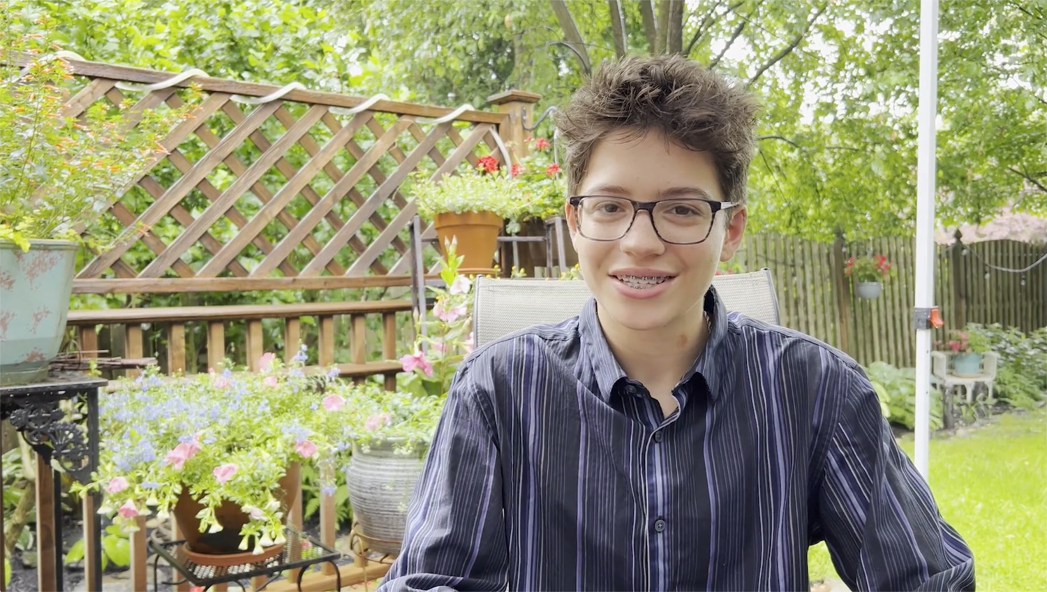 A young man wearing a striped navy blue button down shirt sitting on a wooden deck in a backyard garden.