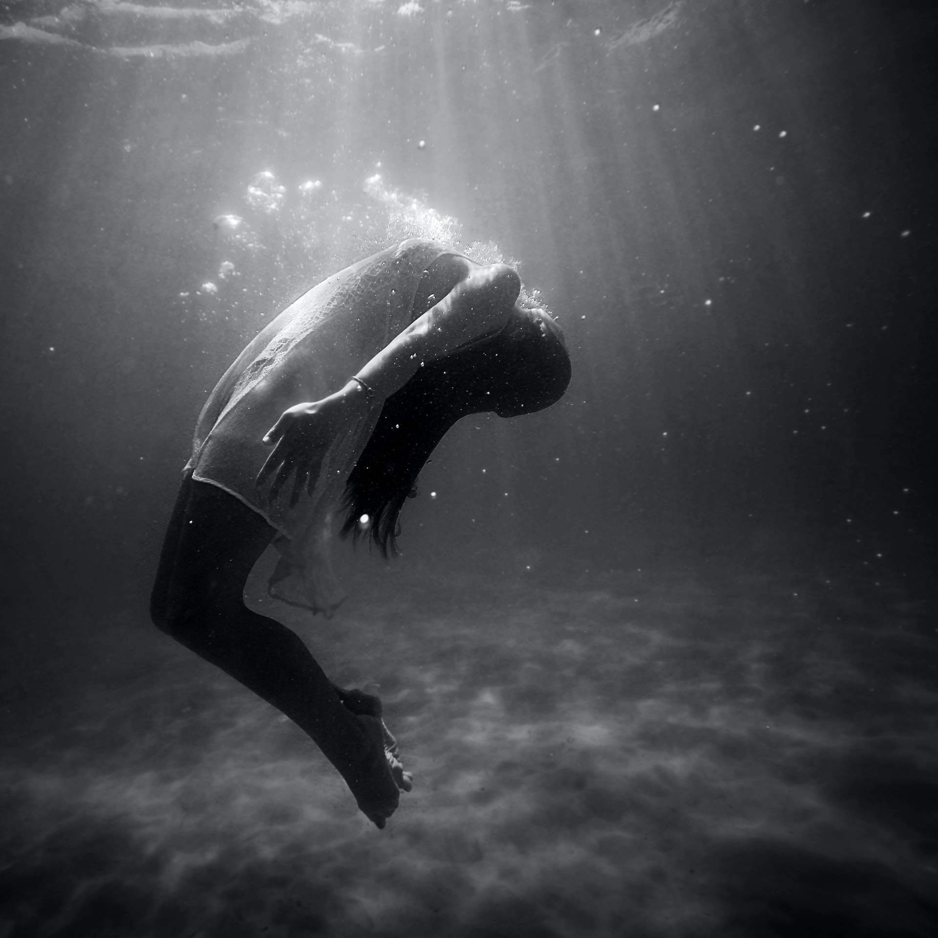greyscale photo of a woman in a white dress underwater with her back arched towards the surface with rays of sunlight shining down