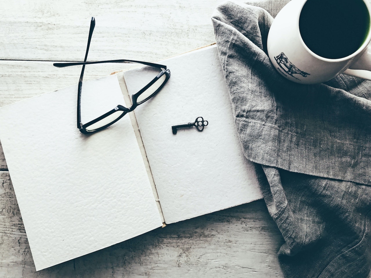 A pair of glasses and a small black key lie on top of a blank notebook. To the right is a grey cloth and a mug.