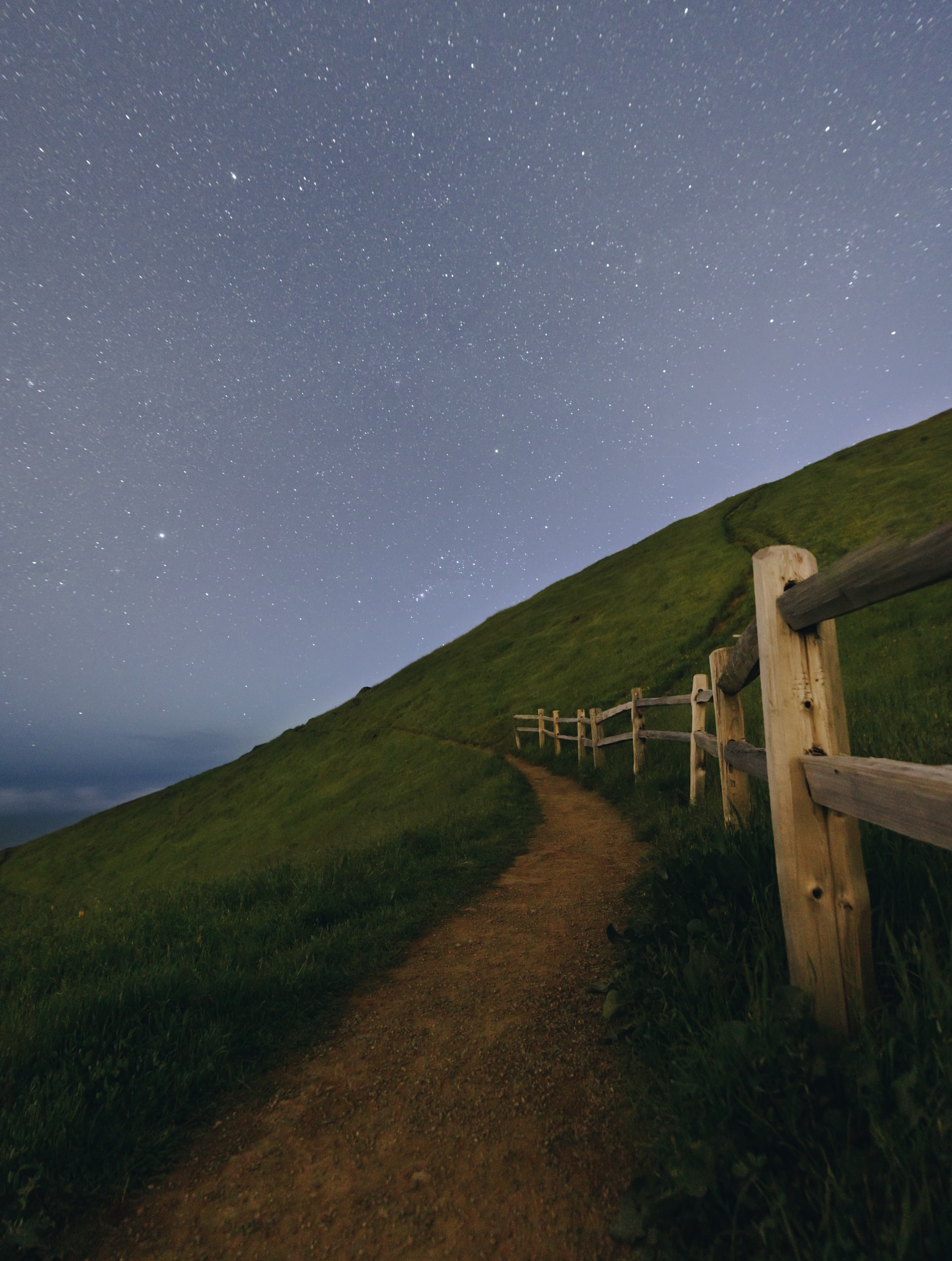 A dirt path winding along a grassy hillside as dusk emerges with stars overhead. The path is bordered on the right hand side by a wooden fence.