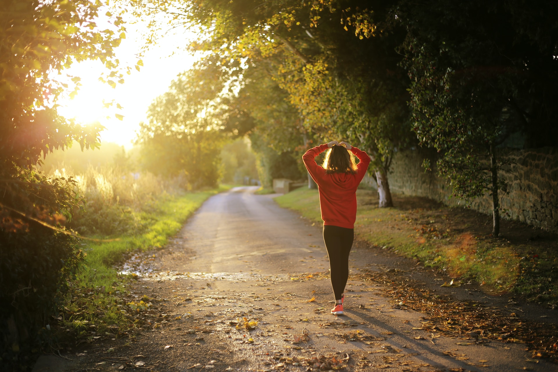 Woman walking down a paved path pausing with her arms over her head to look at the morning sun