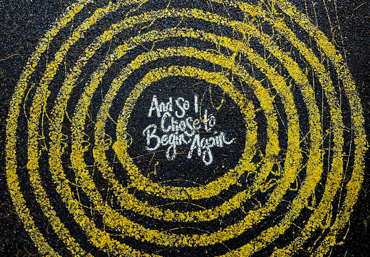 Yellow circles drawn on black pavement with chalk and the words "and so I chose to begin again" in the middle