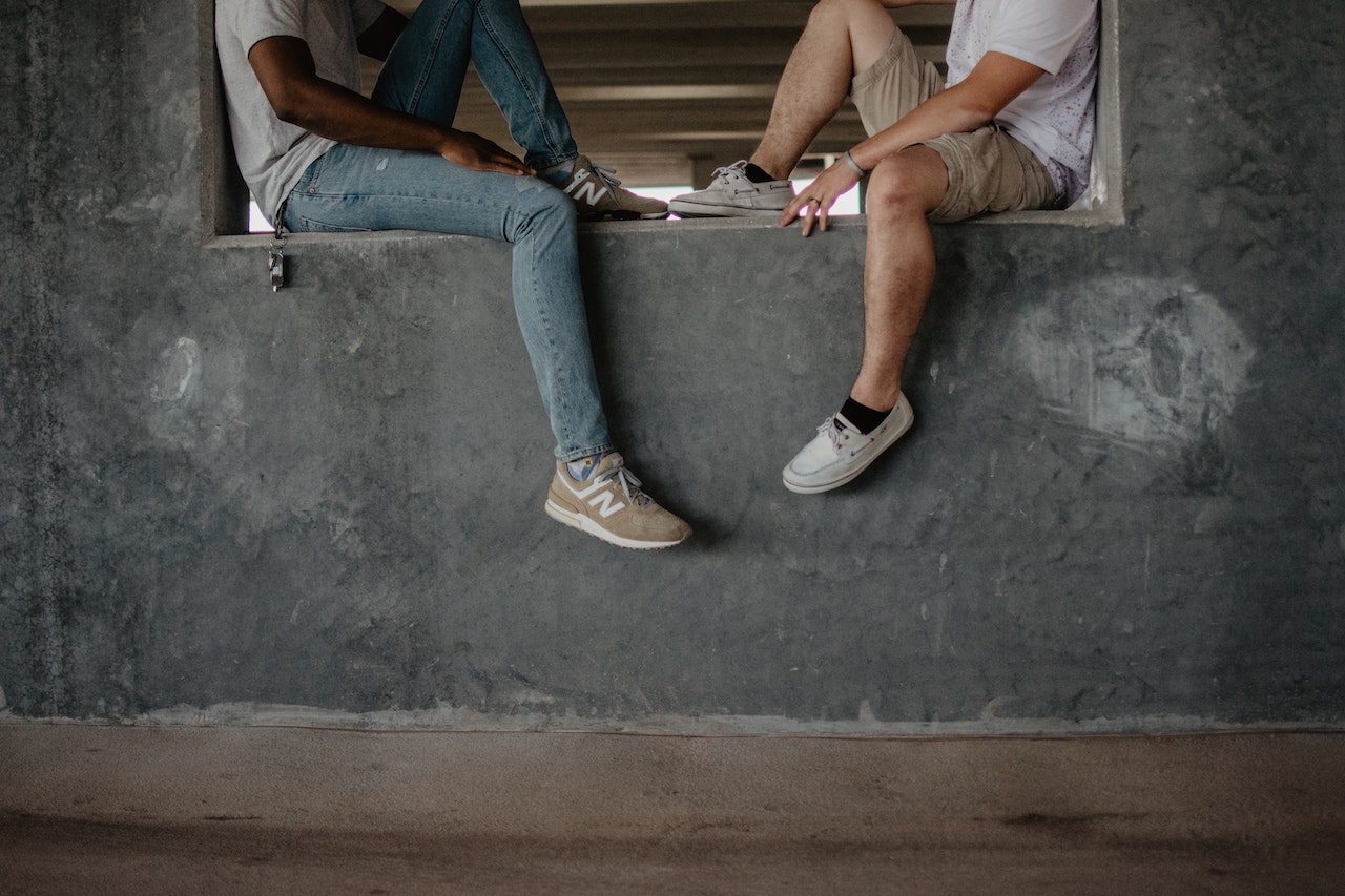 Two people sitting facing each other on a wall. Their bodies are visible from their necks down. One is wearing jeans and the other person is wearing shorts.
