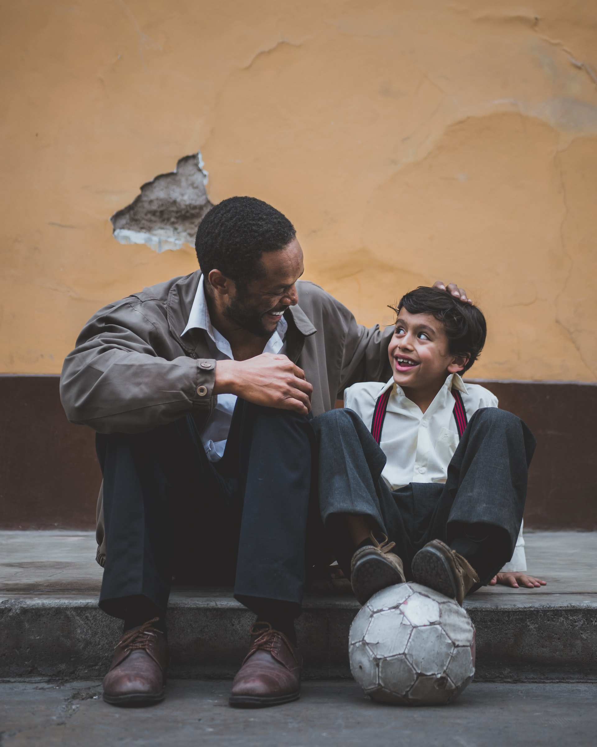 A man and young boy sit next to each other on a curb looking and smiling at each other. The young boy has his feet resting on a soccer ball.