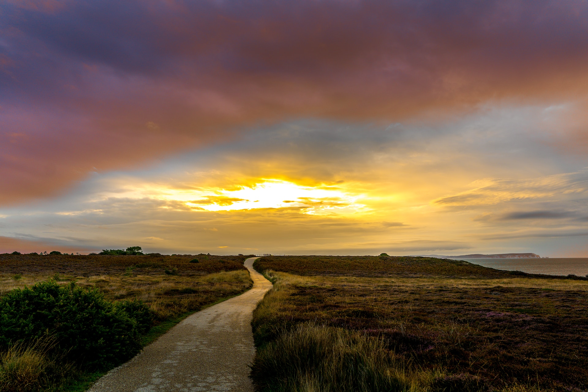 A gently curving gravel pathway surrounded by grasses and water to the right, with the sun rising in the distance