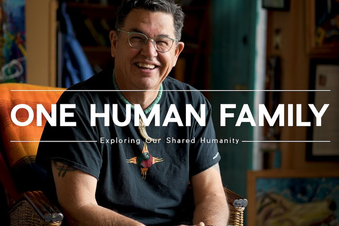 Man sitting on an orange chair smiling with text that reads, "One Human Family"