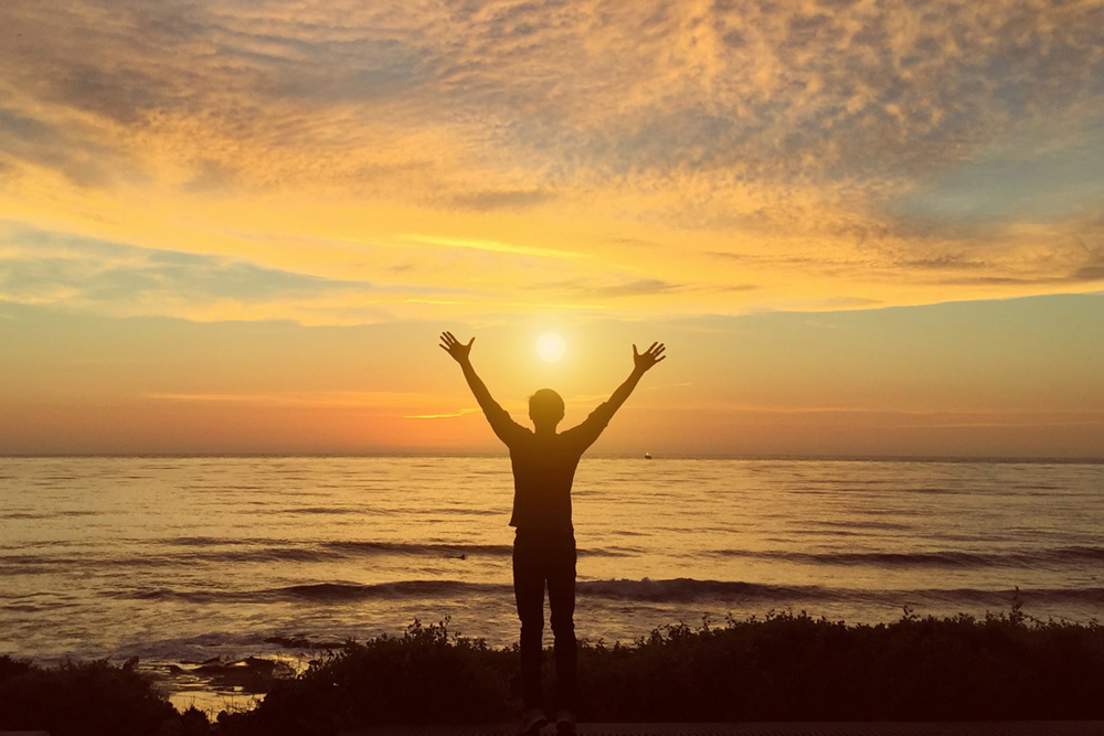 man with arms up silhouetted in front of ocean at sunset