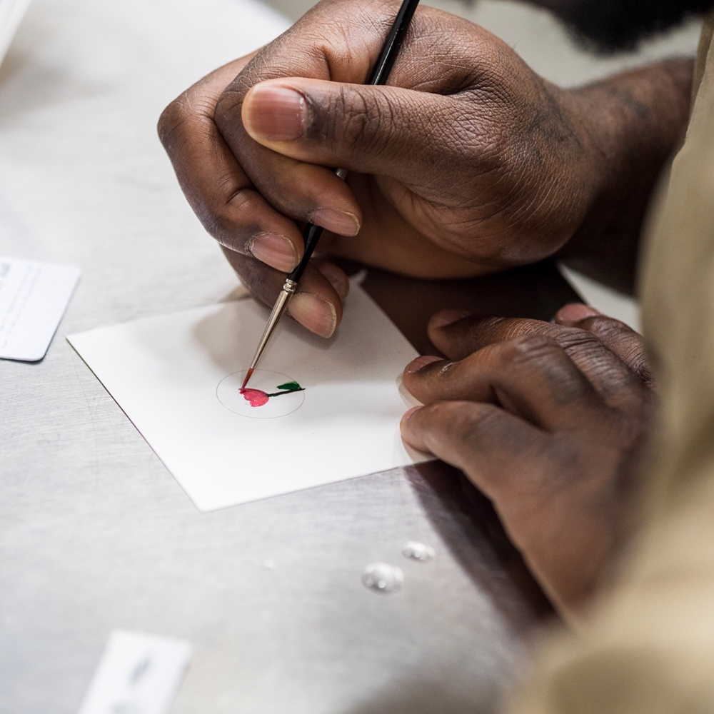 A close up photo of a man's hand painting a small red tulip on a square piece of white paper