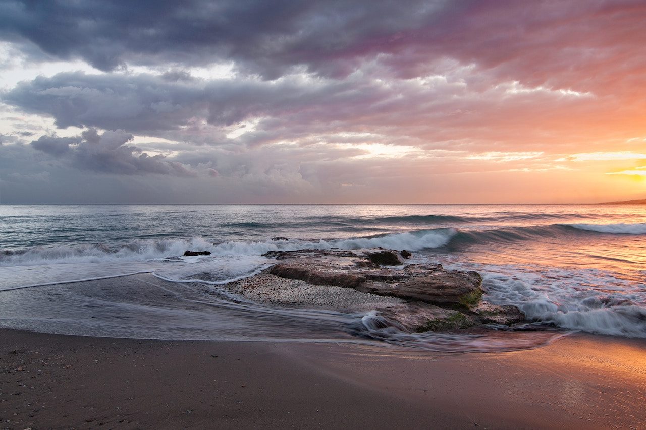 Sunset over a beach with a wave washing over a rock.