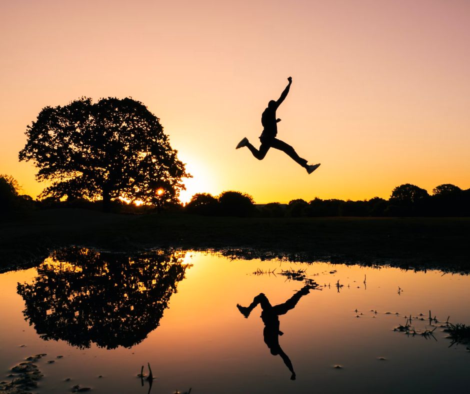 Silhouhette of a man jumping over a body of water with a reflection during golden hour