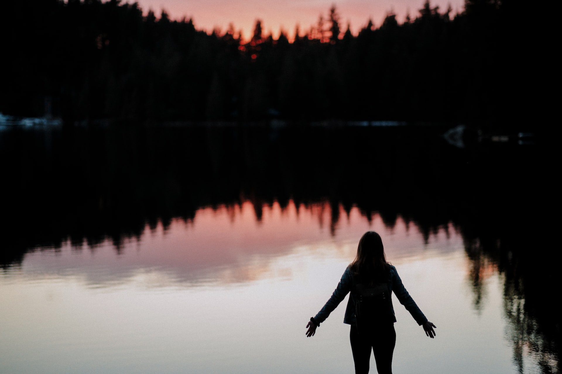 silhouette of a woman standing at the shore of a tree lined lake at sunset with her arms open