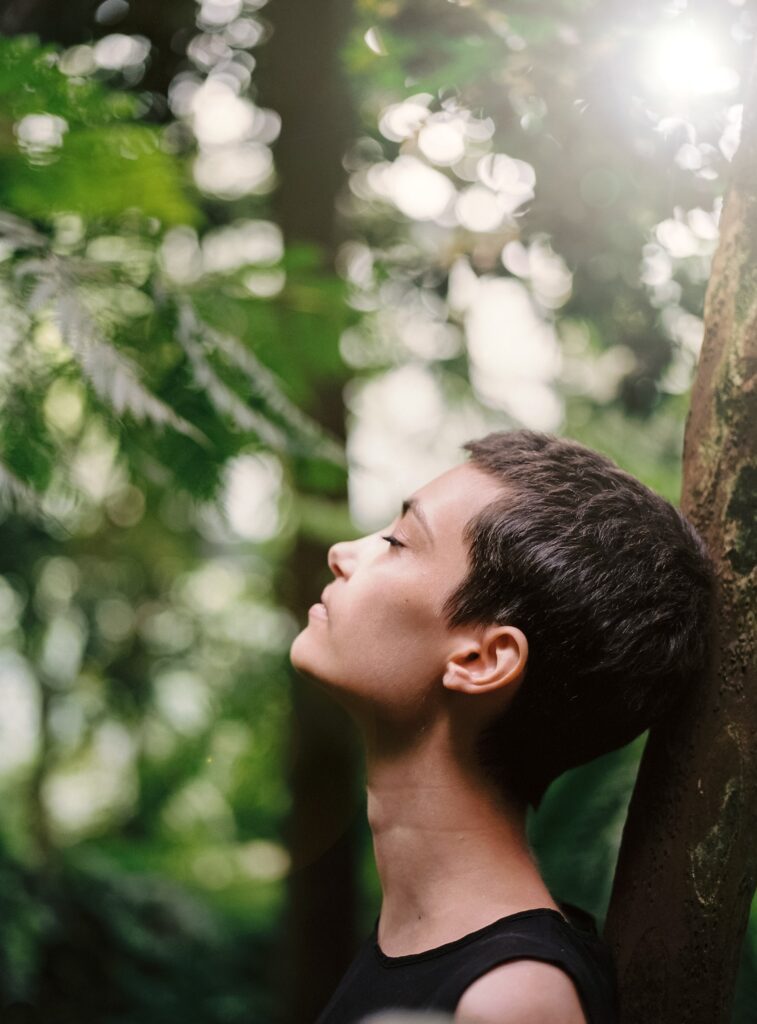 Closeup of a woman with short brown hair calmly resting her head against the trunk of a tree with her eyes closed and head tilted back. Green foliage is blurred in the background.