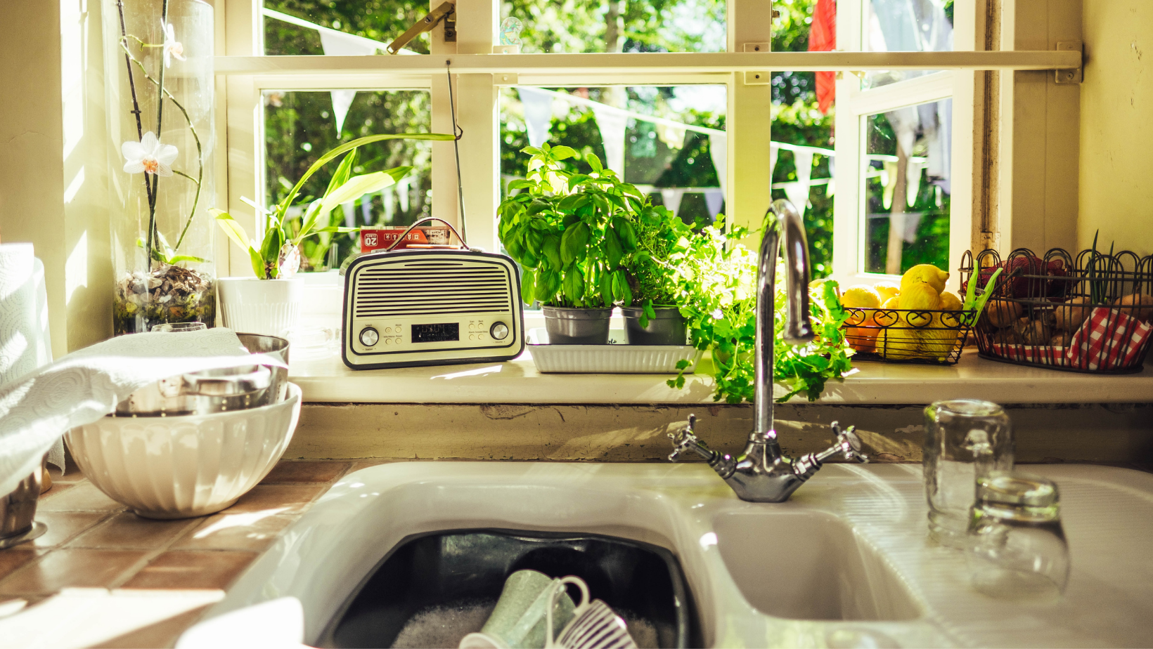 Kitchen sink filled with soaking dishes, up again a sunny window and a window sill with a small radio, herbs, and a fruit bowl.